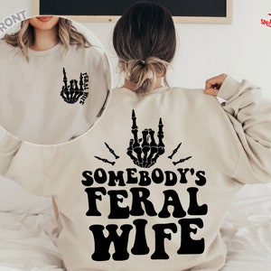 May include: A beige sweatshirt with a black skeleton hand design on the front and the text "Somebody's Feral Wife" on the back.