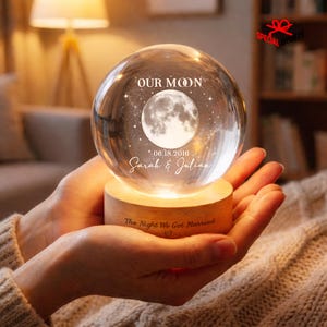 May include: A clear glass sphere resting on a cylindrical light-colored wooden base. The sphere features an etched moon graphic with the text OUR MOON, the date 06.18.2016, and the names Sarah & Julian. The wooden base is engraved with The Night We Got Married.