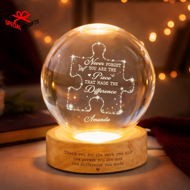 May include: A clear glass sphere lamp resting on a round wooden base. The sphere features an etched puzzle piece design with the text "Never forget you are the piece that made the difference" and the name "Amanda". The base has engraved text.