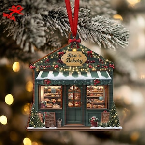 May include: A wooden Christmas ornament shaped like a bakery, with the text "Lisa's Bakery" on a sign. The bakery has a green awning, string lights, and a red ribbon for hanging. The windows display baked goods.
