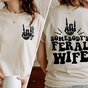 May include: A beige t-shirt with a black skeleton hand making a rock and roll gesture and the text "Feral Wife" on the front. The back of the shirt has the text "Somebody's Feral Wife" in black.