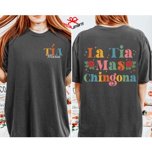 以下が含まれることがあります： 黒いTシャツで、後ろには「La Tia Mas Chingona」というカラフルなグラフィックが描かれており、赤いバラと星が描かれています。Tシャツの前には、「Tia Maria」という小さなグラフィックがカラフルな文字で描かれています。