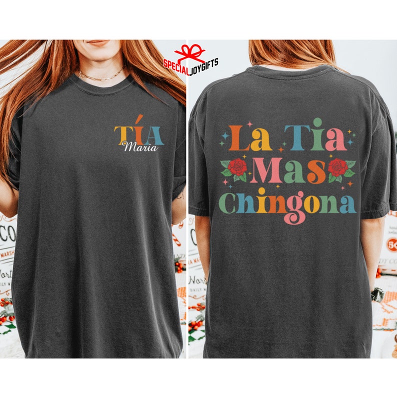 以下が含まれることがあります： 黒いTシャツで、後ろには「La Tia Mas Chingona」というカラフルなグラフィックが描かれており、赤いバラと星が描かれています。Tシャツの前には、「Tia Maria」という小さなグラフィックがカラフルな文字で描かれています。