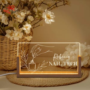 May include: A rectangular LED night light with a wooden base. The light features a nail polish bottle and hand design, with the name "Rebecca" and the words "NAIL TECH" in white script. The light is surrounded by white flowers.