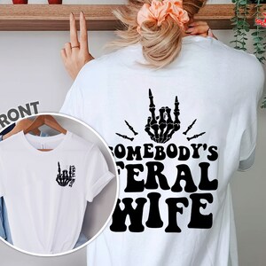 May include: White t-shirt with a black skeleton hand making a rock and roll sign and the text "Somebody's Feral Wife".