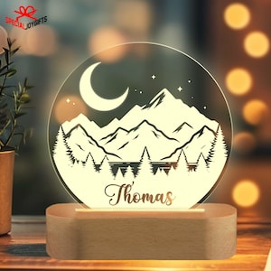 May include: A personalized night light with a mountain scene, a crescent moon, and stars. The name "Thomas" is written below the scene. The light is on a wooden base.