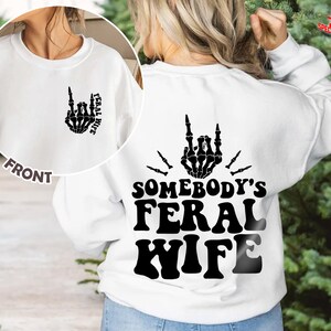 May include: White sweatshirt with a black skeleton hand making a rock and roll sign with the text "Somebody's Feral Wife" on the back. The front of the sweatshirt has a smaller version of the skeleton hand with the text "Feral Wife" on the front.