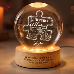 May include: A clear glass sphere lamp resting on a cylindrical wooden base. The sphere features an etched puzzle piece design with the text "Difference Maker THANK YOU FOR BEING SUCH AN IMPORTANT PIECE IN OUR TEAM" and the name "Taylor".