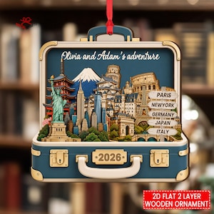 May include: A suitcase-shaped wooden ornament featuring a layered travel scene with the Statue of Liberty, Mount Fuji, and the Colosseum. The text reads Olivia and Adam's adventure, 2026, and a signpost with Paris, Newyork, Germany, Japan, and Italy.