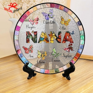 Puede incluir: Un plato redondo de vidrio coloreado con la palabra "NANA" en el centro. Las letras est&aacute;n decoradas con flores y mariposas. El plato est&aacute; rodeado por un borde de piezas de vidrio de colores. Los nombres de ocho nietos est&aacute;n escritos alrededor del plato: Harper, Charlotte, Ellie, Ava, Olivia, Emma, Sophia.