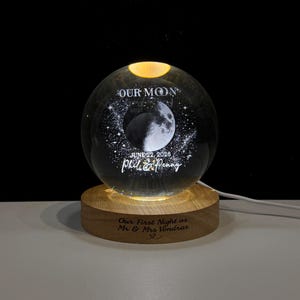May include: A clear glass sphere lamp featuring an internal laser-etched moon and star design. The text reads OUR MOON, JUNE 22, 2026, and Phil & Penny. It sits on a round wooden base engraved with the text Our First Night as Mr & Mrs Vondras.