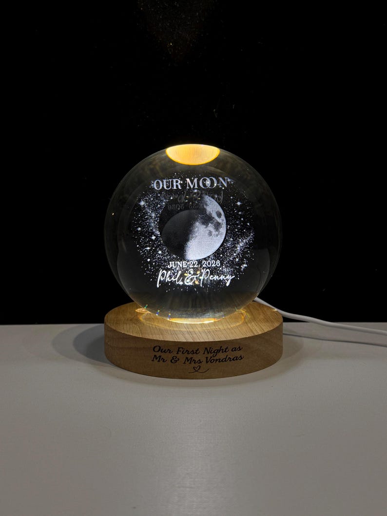 May include: A clear glass sphere lamp featuring an internal laser-etched moon and star design. The text reads OUR MOON, JUNE 22, 2026, and Phil & Penny. It sits on a round wooden base engraved with the text Our First Night as Mr & Mrs Vondras.