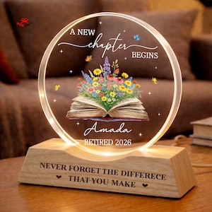 May include: A lighted, circular acrylic plaque with the words "A New Chapter Begins" and "Amada Retired 2026." The plaque features a floral bouquet over an open book and sits on a wooden base with the inscription "Never Forget The Difference That You Make."