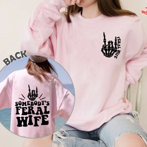 May include: Pink sweatshirt with a black skeleton hand giving the middle finger and the text "Feral Wife" on the front. The back of the sweatshirt has a black skeleton hand giving the middle finger and the text "Somebody's Feral Wife".