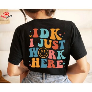 Pu&ograve; includere: Maglietta nera con una grafica in stile retr&ograve; che dice "IDK I JUST WORK HERE" in lettere colorate con una faccina sorridente con fulmini nella parola "WORK".