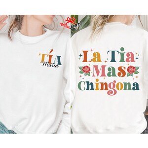 以下が含まれることがあります： 白いスウェットシャツ。背面には「La Tia Mas Chingona」というカラフルなグラフィックが描かれており、花と星が描かれています。スウェットシャツの前面には、「Tia Maria」という小さなグラフィックがあります。