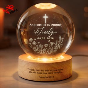 May include: A lighted crystal sphere with a wooden base. The sphere has a cross, the words "CONFIRMED IN CHRIST", the name "Jocelyn", and the date "04.26.2026" engraved on it. The base has the text "Trust in the Lord with all your heart; He will make your paths straight. Proverbs 3:5-6".