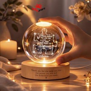May include: A clear glass sphere lamp resting on a round wooden base. The sphere features an etched puzzle piece design with the text "Never forget you are the piece that made the difference" and "Amanda". The base has engraved text.