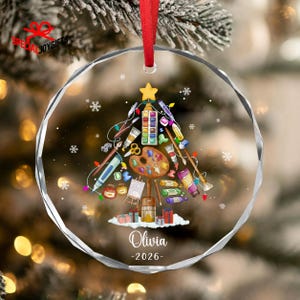 May include: A circular clear acrylic ornament featuring a Christmas tree graphic composed of various art supplies including paintbrushes, a palette, tubes of paint, and pencils. The name Olivia and the year 2026 are printed in white below the tree.