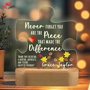 May include: A light-up wooden plaque shaped like a jigsaw piece with the text "Never forget you are the piece that made the difference."  The plaque has a colourful floral design and the name "Grace Taylor" at the bottom.  The plaque is a retirement gift for a mentor, advocate, and friend.