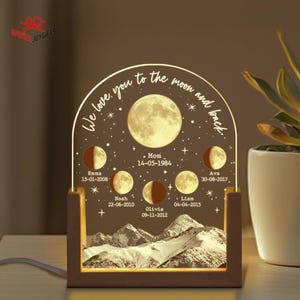 May include: A wooden night light featuring a crescent moon design. The moon is divided into sections, each with a name and date. The text "We love you to the moon and back." is written around the moon. The night light is illuminated with a warm glow.