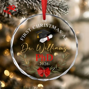 May include: A round, faceted clear glass ornament hanging from a red ribbon. It features the text "FIRST CHRISTMAS AS Dr. Williams PhD -2026-" in black and red, with a black graduation cap, a white diploma, and a green wreath with a red bow.