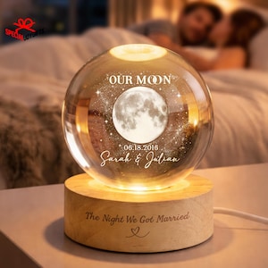 May include: A spherical glass orb featuring an etched moon and star design, the text "OUR MOON", the date "06.18.2016", and the names "Sarah & Julian". The orb rests on a cylindrical wooden base engraved with "The Night We Got Married" and a heart.