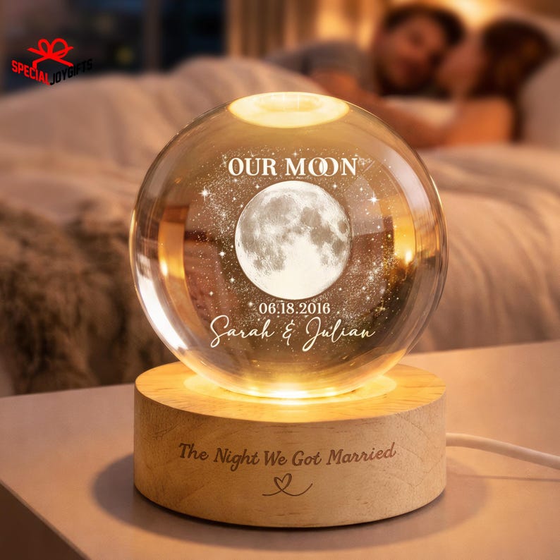 May include: A spherical glass orb featuring an etched moon and star design, the text "OUR MOON", the date "06.18.2016", and the names "Sarah & Julian". The orb rests on a cylindrical wooden base engraved with "The Night We Got Married" and a heart.