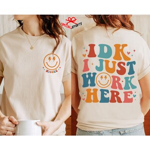 Pu&ograve; includere: Una maglietta beige con un viso sorridente con fulmini e il nome "Nicole" sul davanti. La parte posteriore della maglietta ha il testo "IDK I JUST WORK HERE" in lettere retr&ograve; colorate con un viso sorridente con fulmini al centro.