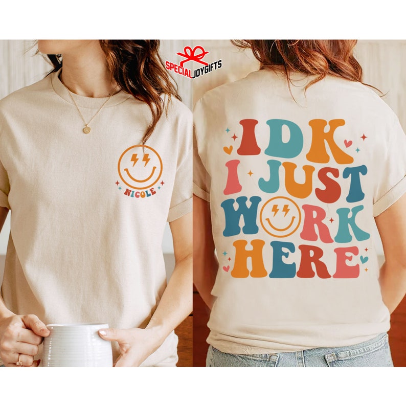 Pu&ograve; includere: Una maglietta beige con un viso sorridente con fulmini e il nome "Nicole" sul davanti. La parte posteriore della maglietta ha il testo "IDK I JUST WORK HERE" in lettere retr&ograve; colorate con un viso sorridente con fulmini al centro.