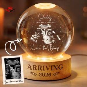 May include: A clear glass sphere lamp on a round wooden base. The sphere features an etched ultrasound image with the text Daddy I can't wait to meet you! Love, the Bump. The wooden base is engraved with ARRIVING 2026 and leaf designs.