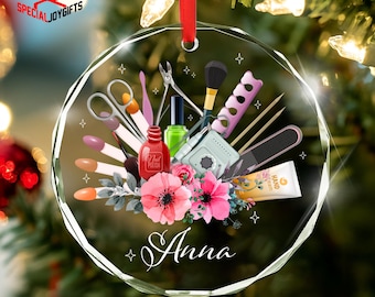 May include: A clear, round Christmas ornament with nail care tools and pink flowers. The ornament features nail polish, scissors, a brush, and the name "Anna" in script. A red ribbon is attached for hanging.
