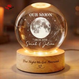 May include: A spherical glass orb featuring an etched moon and star design, with the text OUR MOON, 06.18.2016, and Sarah & Julian. The orb rests on a cylindrical light-colored wooden base engraved with The Night We Got Married and a heart symbol.
