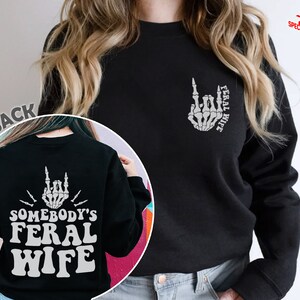 May include: Black sweatshirt with a white skeleton hand making a middle finger gesture and the text "Feral Wife" on the back. The front of the sweatshirt has the text "Feral Wife" on the left chest.