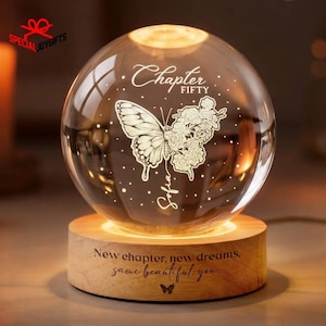 May include: A clear glass sphere with a butterfly and floral design, illuminated from within, sits on a wooden base. The sphere has the words "Chapter Fifty" and "Sofia" etched on it. The base has the text "New chapter, new dreams, same beautiful you."