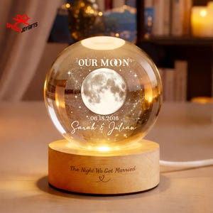 May include: A clear glass sphere lamp featuring an etched moon and star design with the text "OUR MOON", "06.18.2016", and "Sarah & Julian". The sphere rests on a cylindrical light-colored wooden base engraved with "The Night We Got Married" and a heart.