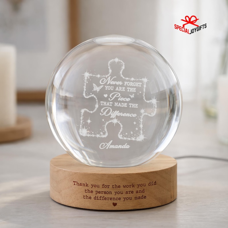 May include: A clear glass sphere resting on a round wooden base. The sphere features an etched puzzle piece design with the text: Never forget you are the piece that made the difference, and the name Amanda. The base has engraved text.