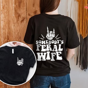 May include: Black t-shirt with a white graphic of a skeleton hand making a rock and roll sign with the text "Somebody's Feral Wife".