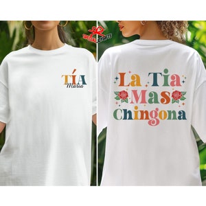 以下が含まれることがあります： 白地に「T&iacute;a Maria」の文字が書かれたTシャツ。後ろにはカラフルな花柄と「La T&iacute;a Mas Chingona」の文字が書かれている。