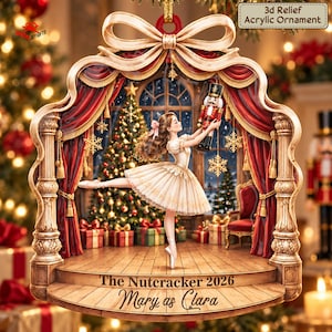 May include: A 3D relief acrylic ornament featuring a ballerina in a white dress holding a nutcracker on a stage with red curtains. The base is inscribed with 'The Nutcracker 2026' and 'Mary as Clara'. A gold bow sits at the top of the frame.