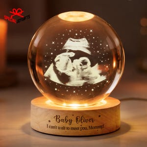 May include: A glowing crystal sphere with an ultrasound image of a baby, set on a wooden base. The base is engraved with "Baby Oliver" and a message. The sphere is illuminated with warm light, creating a soft, inviting glow.