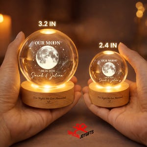 May include: Two clear glass crystal spheres on light wood bases, illuminated from below. The larger sphere is 3.2 inches, the smaller is 2.4 inches. Both feature a moon engraving, the text "OUR MOON", "06.18.2016", "Sarah & Julian", and "The Night We Got Married".