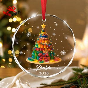 May include: A circular clear glass ornament featuring a printed graphic of a Christmas tree constructed from colorful pencils and paint pots. The tree is topped with a yellow star, adorned with a red plaid bow, and includes the name Laura and 2026.