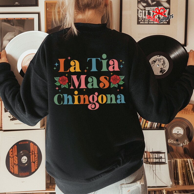 以下が含まれることがあります： 赤いバラと星が描かれた「La Tia Mas Chingona」というカラフルなグラフィックがプリントされた黒いスウェットシャツです。