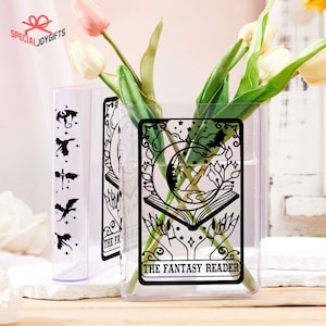 May include: Two clear glass vases with black graphic designs. The vase on the left has a dragon, a bird, a cross, and a bird design. The vase on the right has a crescent moon, a book, mountains, and stars. The text "THE FANTASY READER" is printed on the vase.