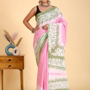 May include: A pink saree with a green and white tie-dye pattern. The saree is draped over a person. The saree has a solid pink base with a green and white patterned border. The person is wearing a black top.