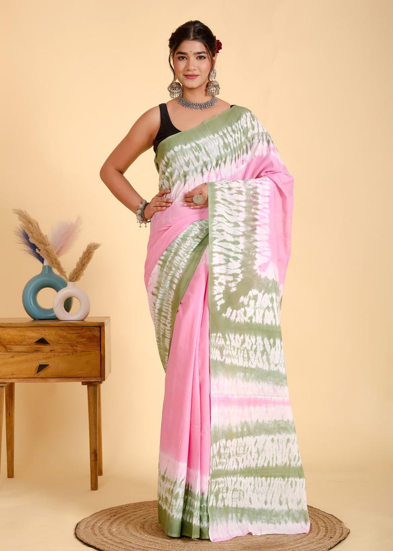 May include: A pink saree with a green and white tie-dye pattern. The saree is draped over a person. The saree has a solid pink base with a green and white patterned border. The person is wearing a black top.