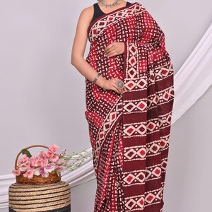 May include: A maroon and white patterned saree. The saree features a repeating geometric design with a border. The model is wearing a black top and silver jewelry. A small basket of pink flowers sits nearby.