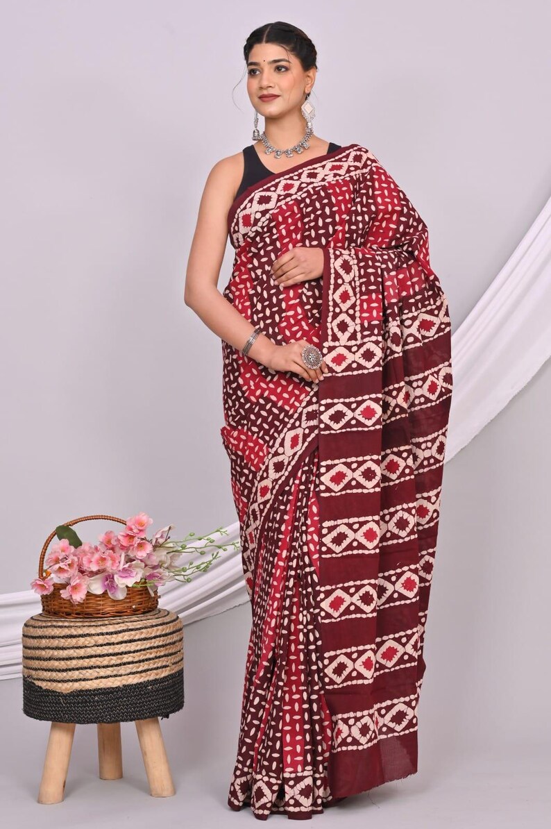 May include: A maroon and white patterned saree. The saree features a repeating geometric design with a border. The model is wearing a black top and silver jewelry. A small basket of pink flowers sits nearby.