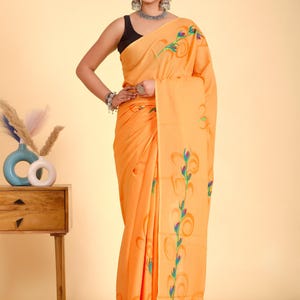 May include: A woman wearing an orange saree with a floral design in green, blue, and orange. The saree is draped over the woman's body. She is wearing a black top and silver jewelry. The background is a light orange color.